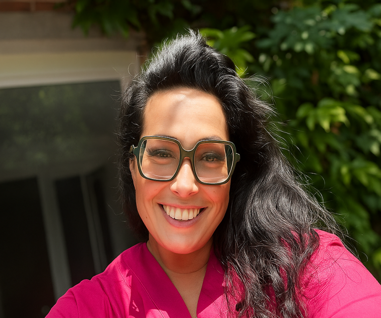 Frau mit langen, schwarzen Haaren und Brille, lächelt vor grüner Pflanzenkulisse.